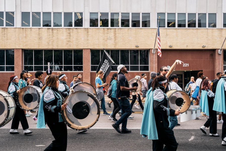 Marching band members invite a wave of energy as their music bounces off the surrounding buildings. A black and white flag reading Black is Beautiful flows above them all. 